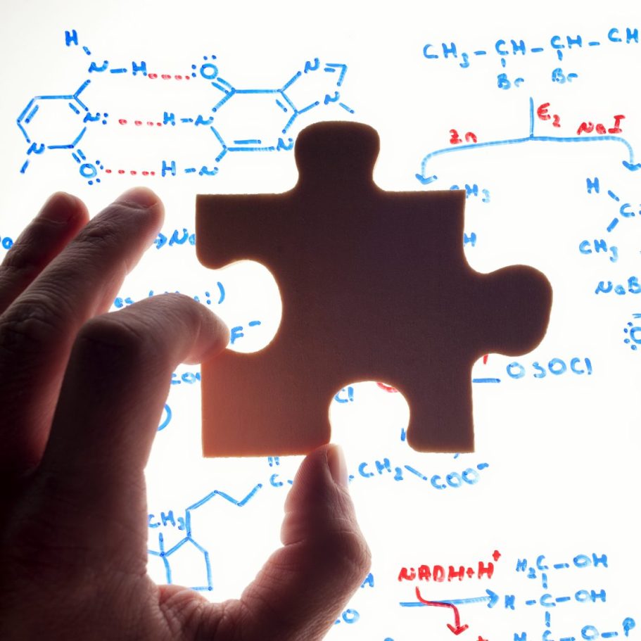 Foto von einem Puzzelstück, welches eine Hand vor eine Tafel mit chemischen Formeln hält