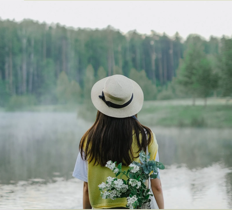 Foto einer Frau mit Hut und Blumen in der Tasche von hinten, da sie übers Wasser schaut und nicht in die Kamera